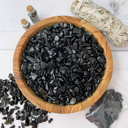 A wooden bowl filled with polished black stones, including Obsidian Crystal Chips, is surrounded by scattered stones, a bundle of dried sage, a small plastic bag of stones, and two small corked glass bottles. The items are arranged on a white surface, creating a minimalist and natural aesthetic.