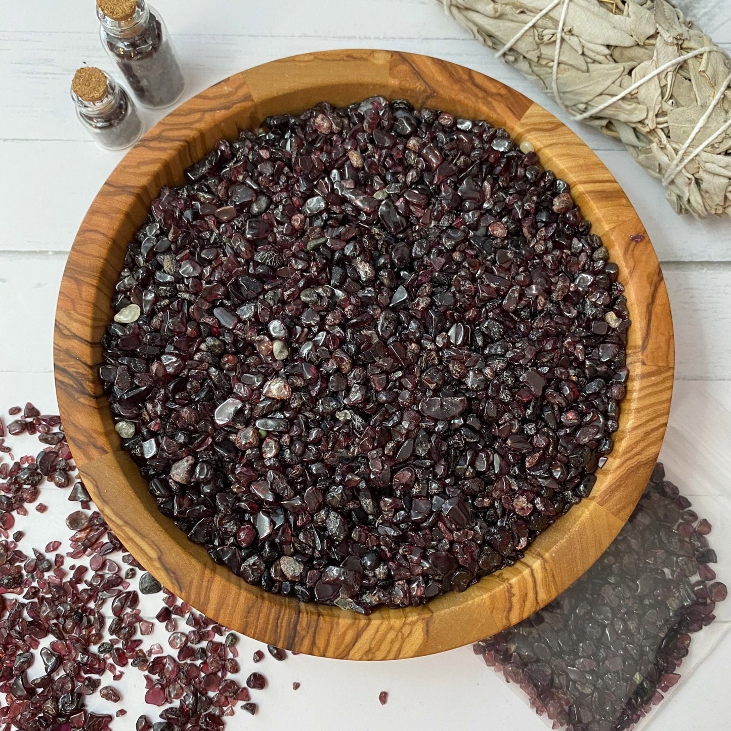 A wooden bowl filled with small, polished Garnet Crystal Chips is centered on a white surface. Surrounding items include a bundle of white sage, two small spell jars, and a partially visible pouch of tumbled and polished stones. Some stones are scattered outside the bowl.