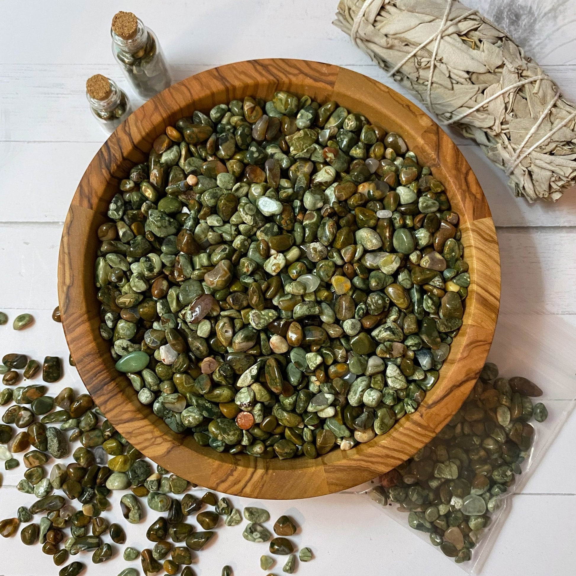 A wooden bowl filled with Rainforest Jasper Crystal Chips is placed on a white surface. Some stones have spilled out of the bowl onto the surface. Nearby, there are two small glass bottles with cork stoppers and a bundle of dried sage wrapped with string.