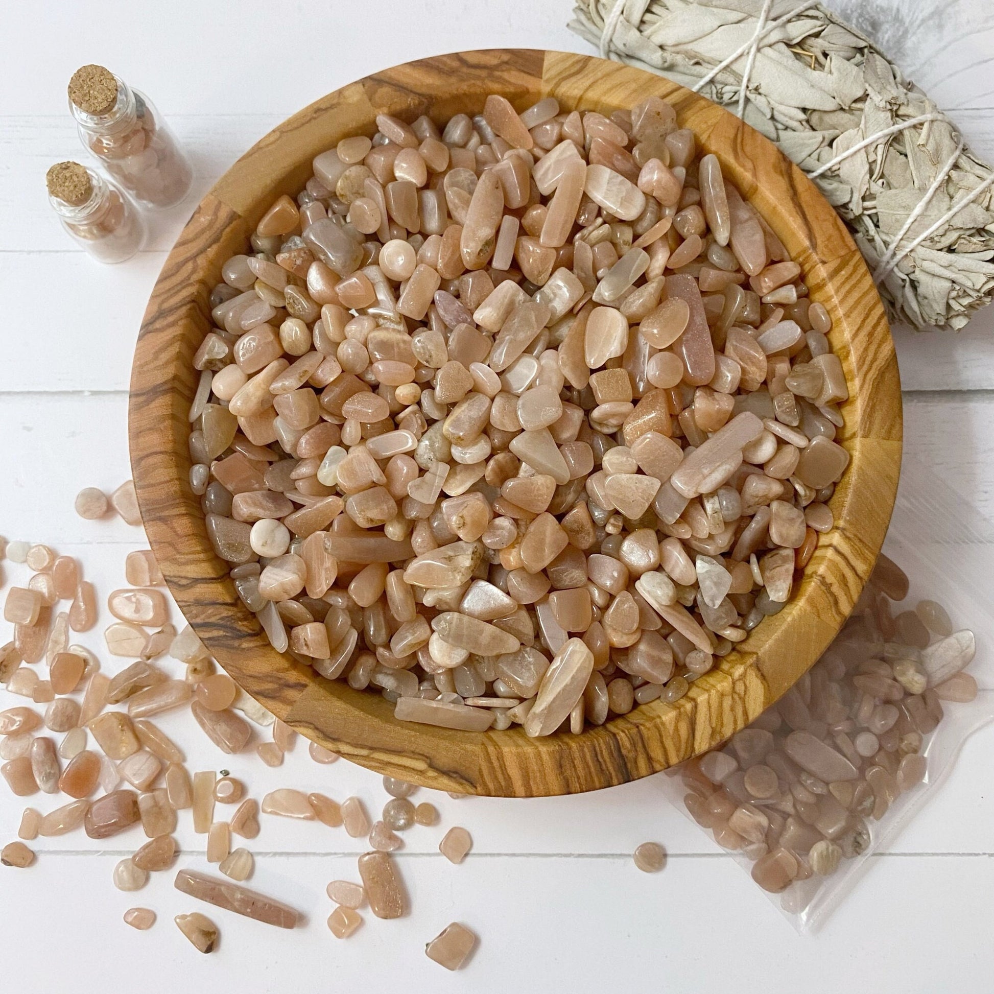 A wooden bowl filled with small peach-colored stones, known as Peach Moonstone Crystal Chips, is centered on a white surface. To the left, some crystal chips are scattered, and two small jars filled with the same stones are placed. A bundle of white sage tied with string is on the top right.