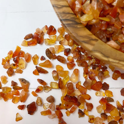 A wooden bowl filled with small, polished Carnelian Crystal Chips in shades of orange and red is centered on a white surface. A smudge bundle, two small vials of stones, and a scatter of additional Carnelian Crystal Chips surround the bowl, perfect for creative and mystical endeavors.