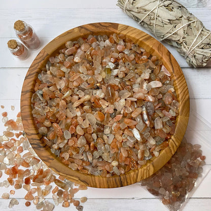 A wooden bowl filled with polished orange and clear crystals, including Sunstone Crystal Chips, sits on a white surface. Scattered crystals surround the bowl, alongside two small spell jars filled with crystals and a bundle of dried sage.