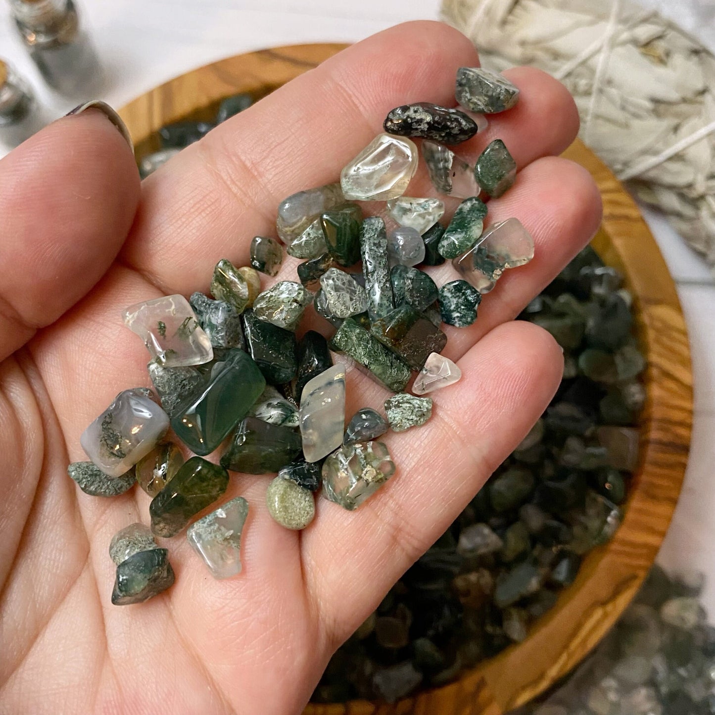 A wooden bowl filled with polished green and clear stones, including green moss agate, sits on a white surface. Beside it are two small vials with cork stoppers, a bundle of dried sage, and a small pile of Green Moss Agate Crystal Chips spilling out of a plastic bag.