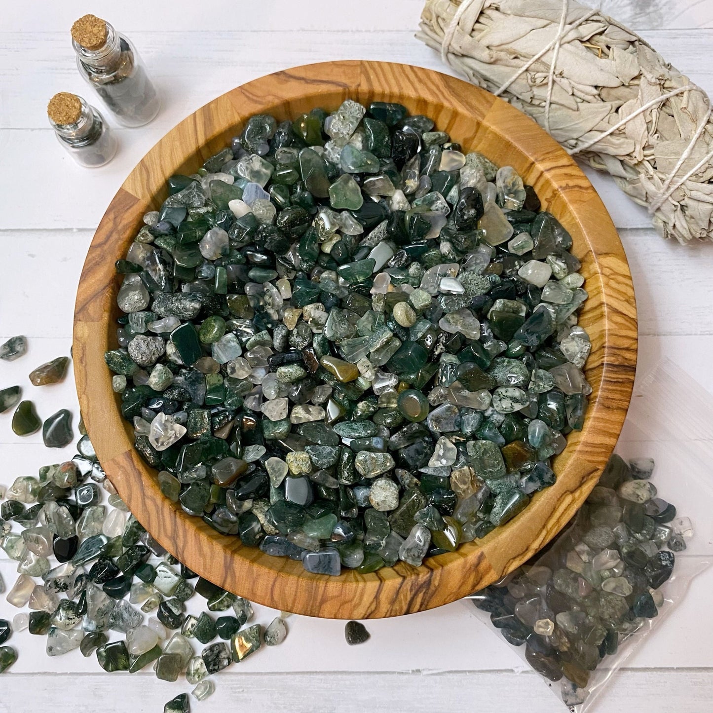 A wooden bowl filled with polished green and clear stones, including green moss agate, sits on a white surface. Beside it are two small vials with cork stoppers, a bundle of dried sage, and a small pile of Green Moss Agate Crystal Chips spilling out of a plastic bag.