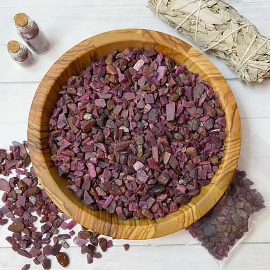 A wooden bowl filled with tumbled tiny stones sits on a white surface, with some purple stones scattered around it. Nearby are two small vials, another small bag of the same stones, and a white sage smudge stick, suggesting metaphysical uses for these Ruby Crystal Chips.