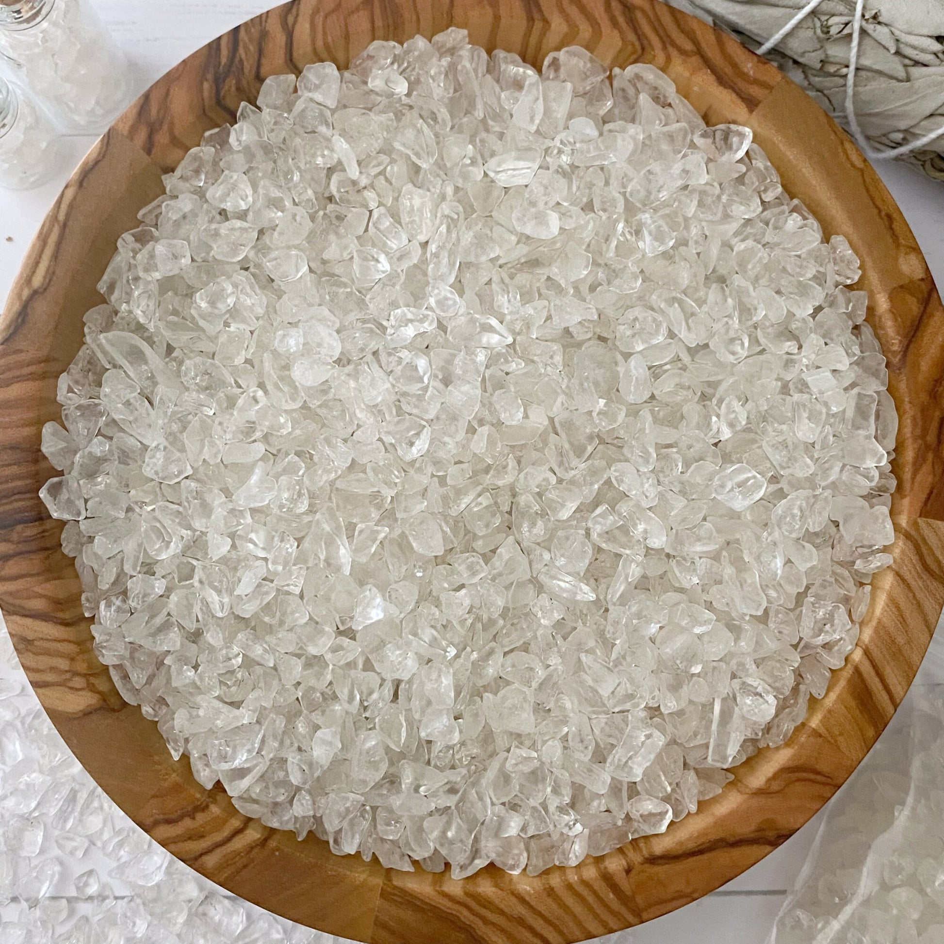 A wooden bowl filled with Clear Quartz Crystal Chips rests on a white surface. Nearby, two small glass bottles with cork stoppers, a bundle of sage tied with string, and a plastic bag partially filled with similar crystal chips complete the serene setting.