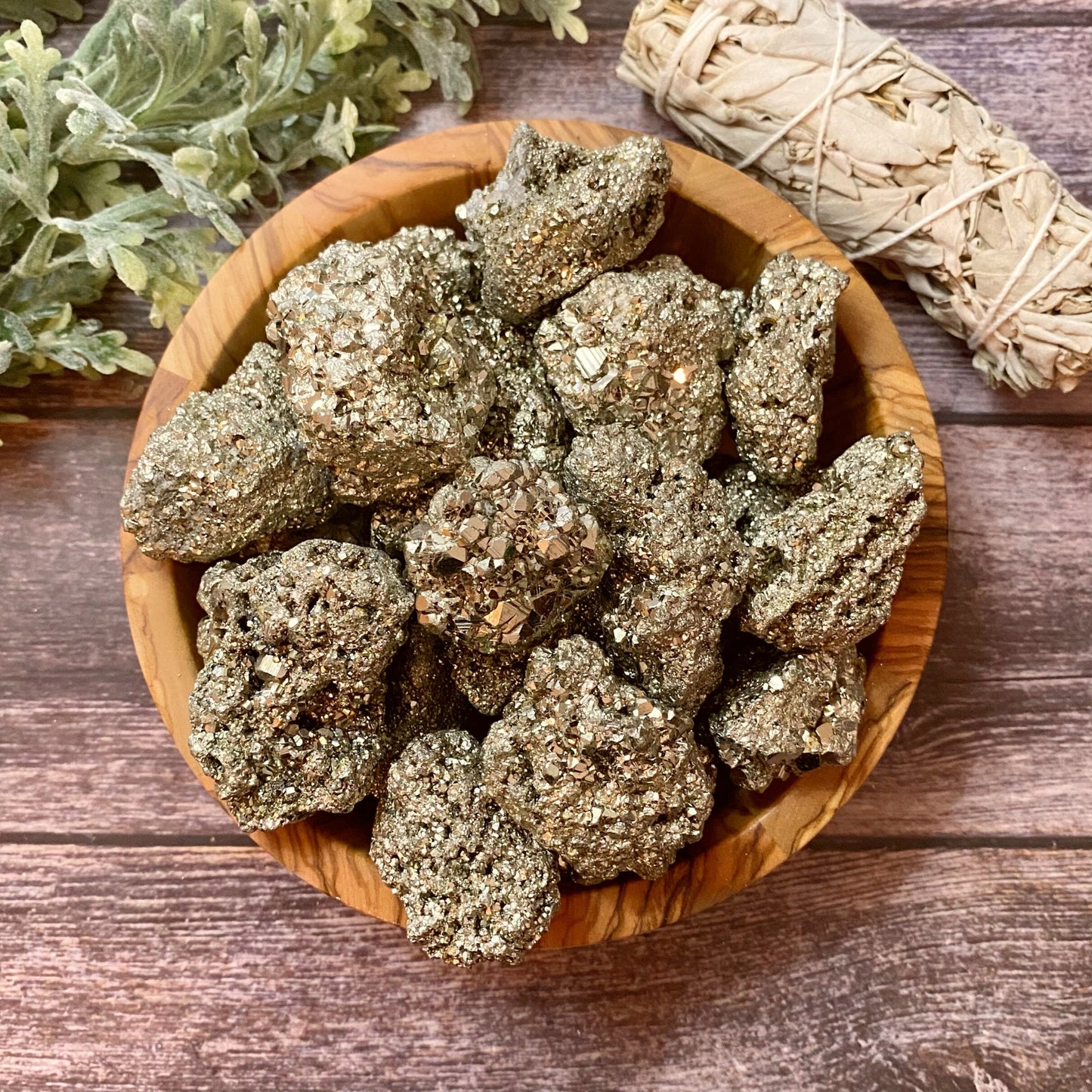 A wooden bowl filled with Raw Pyrite Stones sits on a wooden surface, perfect for meditation. Beside the bowl, there is a bundle of sage wrapped in twine. Lush green leaves are partially visible in the background.