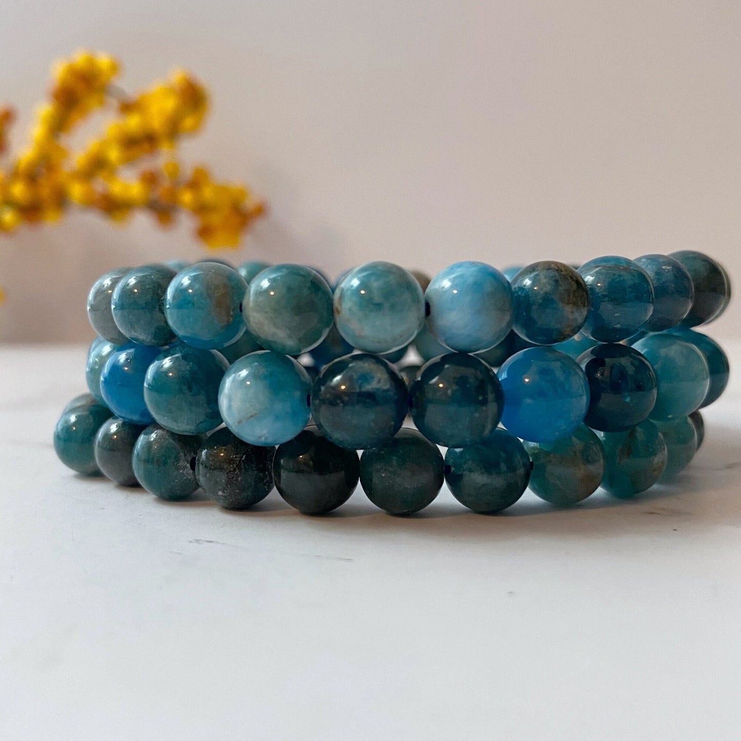 Three stacked 8mm Apatite Beaded Bracelets made of round beads in varying shades of blue, from light to dark. The bracelets are laid out on a white surface with some small yellow flowers blurred in the background, enhancing their connection to spiritual attunement and psychic gifts.