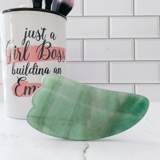 A Green Aventurine Gua Sha is in the foreground on a white surface. In the background, there's a white mug with the text "just a Girl Boss building an Empire," partially covered by a pink stripe. The mug contains various makeup brushes against a white tiled wall.