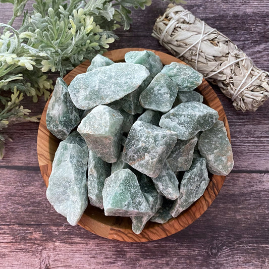 A wooden bowl filled with Raw Green Aventurine Stones rests on a rustic wooden surface. Nearby, a bundle of sage is tied together with string, and some leafy green plants are visible in the background, creating a natural and earthy setting that evokes prosperity and wealth.