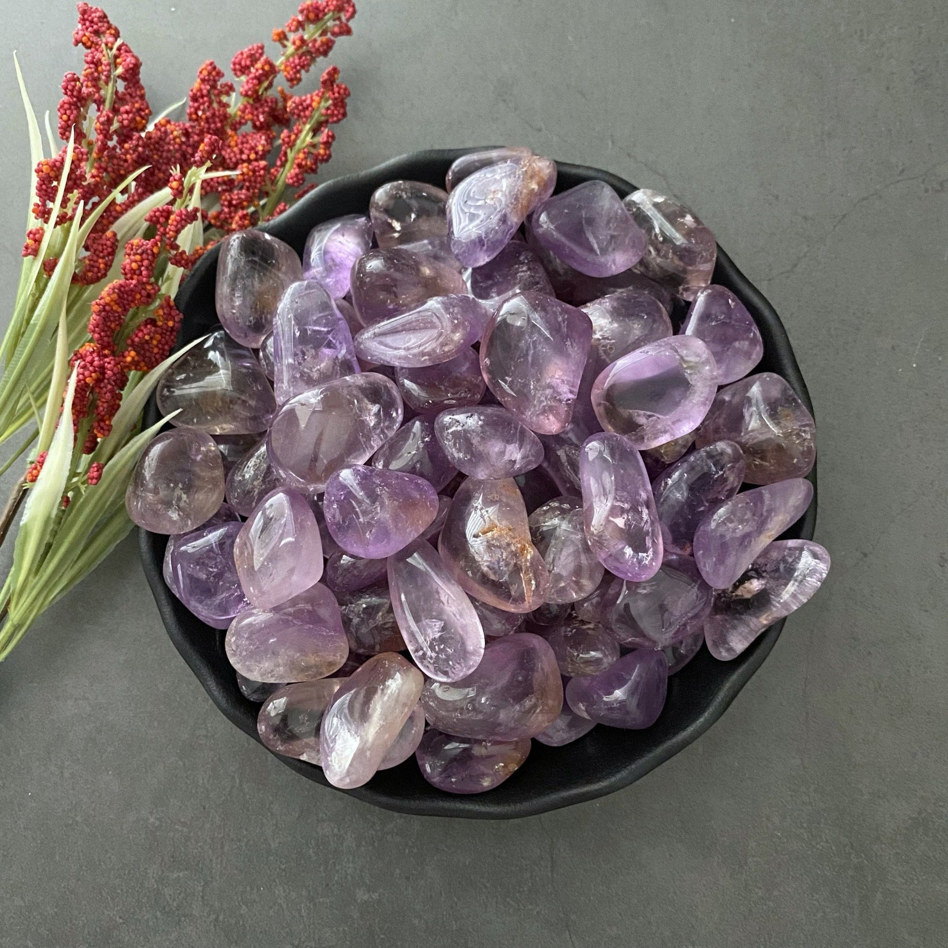 A black bowl filled with Ametrine Tumbled Stones, ranging in shades of purple and yellow, sits on a dark surface. To the left of the bowl, sprigs of red berries and green foliage add a touch of natural color to this calming composition perfect for stress relief.