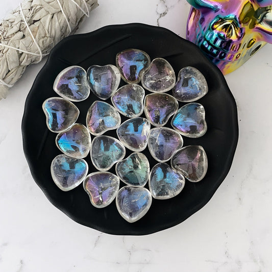 A black dish holds multiple 1" Mini Angel Aura Crystal Quartz Heart stones. The dish is placed on a white marble surface. Nearby, there is a bundle of sage and a colorful, metallic skull.