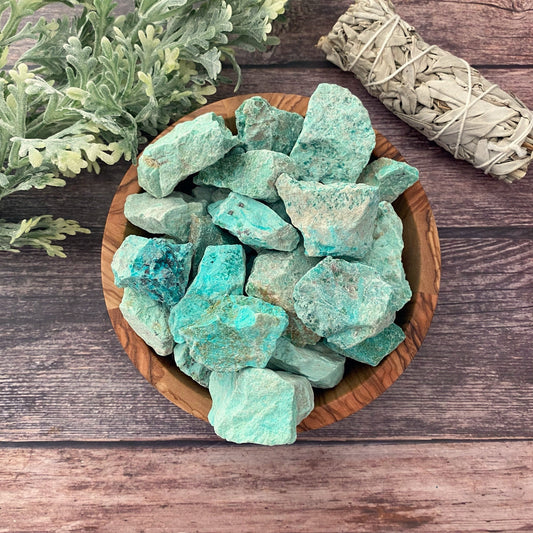 A wooden bowl filled with Raw Peruvian Turquoise Stones sits on a wooden surface. Next to the bowl, there is a bundle of dried sage and sprigs of green foliage. The scenery creates a natural and earthy ambiance, evoking the powerful essence of purification stones.