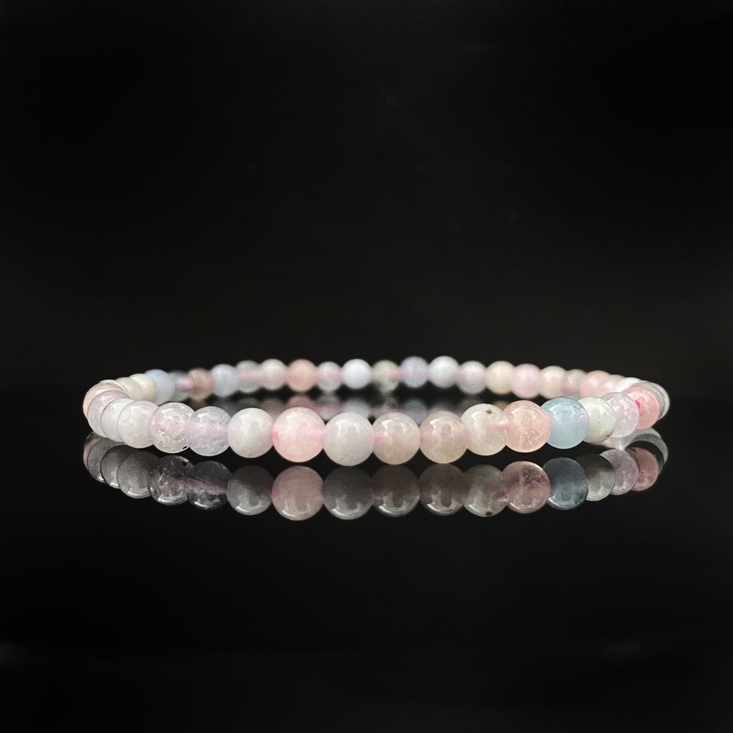 A delicate 4mm Morganite Beaded Bracelet with round pastel beads in shades of pink, white, and light blue, set against a black background. The bracelet is neatly arranged in a circle and reflects on the glossy black surface beneath it, ideal for meditation healing.