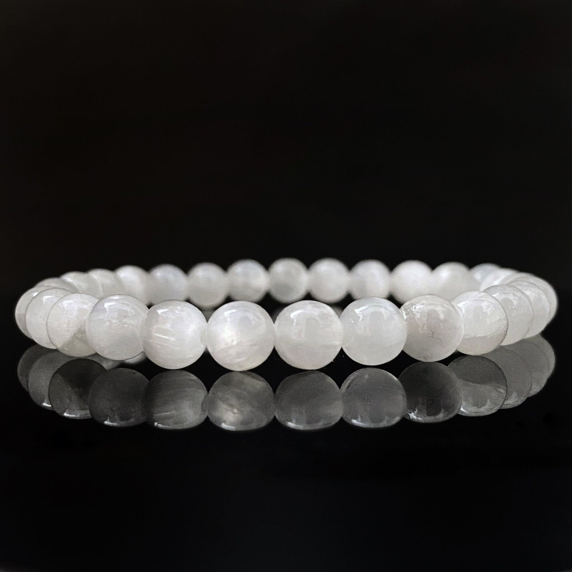 A close-up image of an 8mm Selenite Beaded Bracelet made from round stones, displayed on a reflective black surface. The bracelet forms a complete circle, with each bead evenly spaced, and the reflective surface creating a mirror effect below.