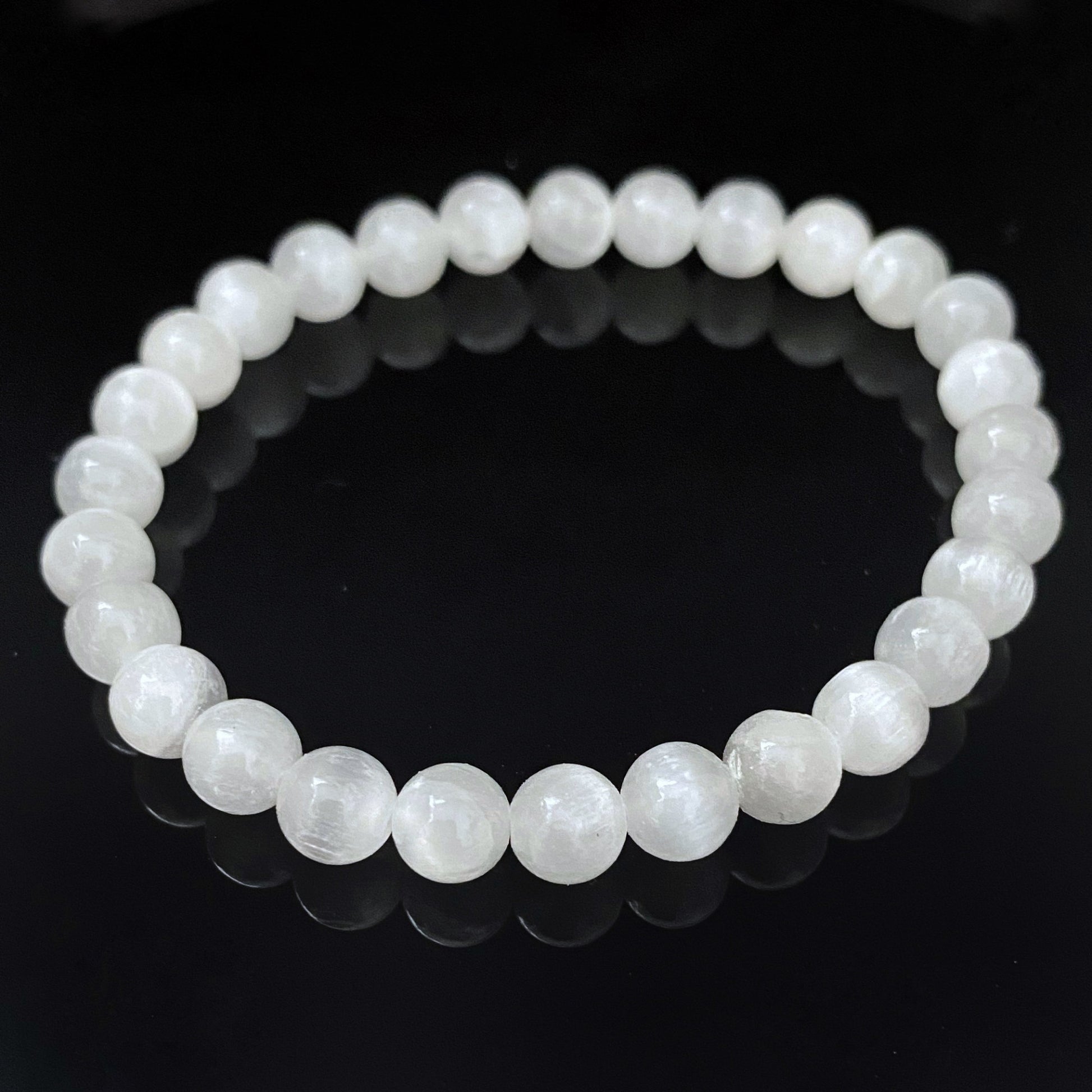 A close-up image of an 8mm Selenite Beaded Bracelet made from round stones, displayed on a reflective black surface. The bracelet forms a complete circle, with each bead evenly spaced, and the reflective surface creating a mirror effect below.