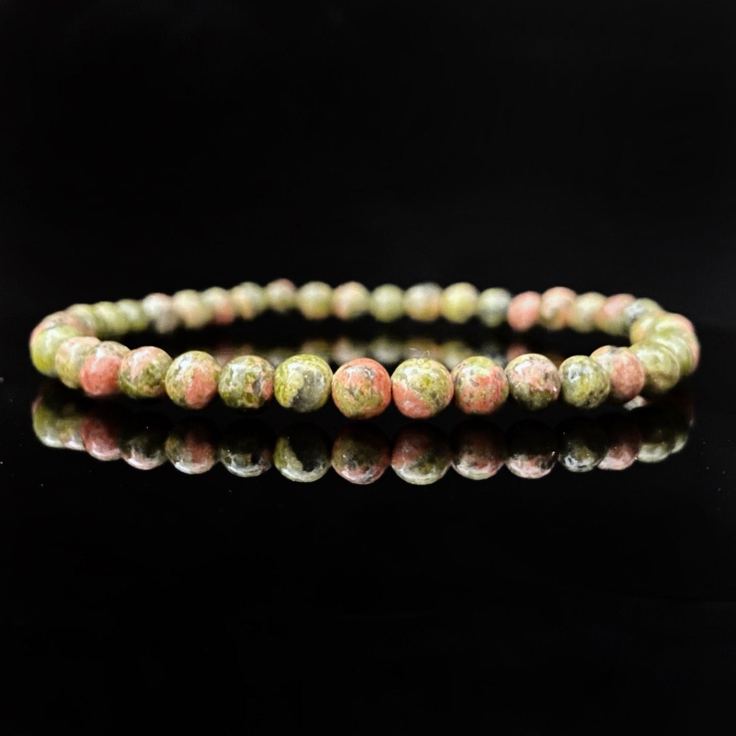 A 4mm Unakite Beaded Bracelet made of round, multicolored stones in shades of green and pink is displayed against a glossy black background, creating a reflection underneath.