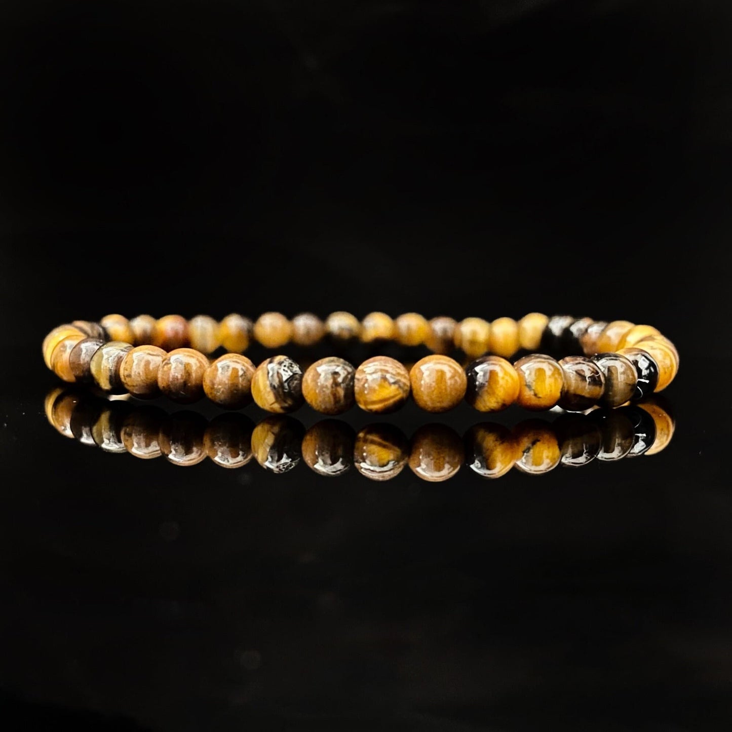 A 4mm Tigers Eye Beaded Bracelet, made of natural stone with spherical beads in golden yellow and brown hues, lies coiled on a shiny black surface. The reflective surface enhances the glossy appearance of the Reiki-infused bracelet.