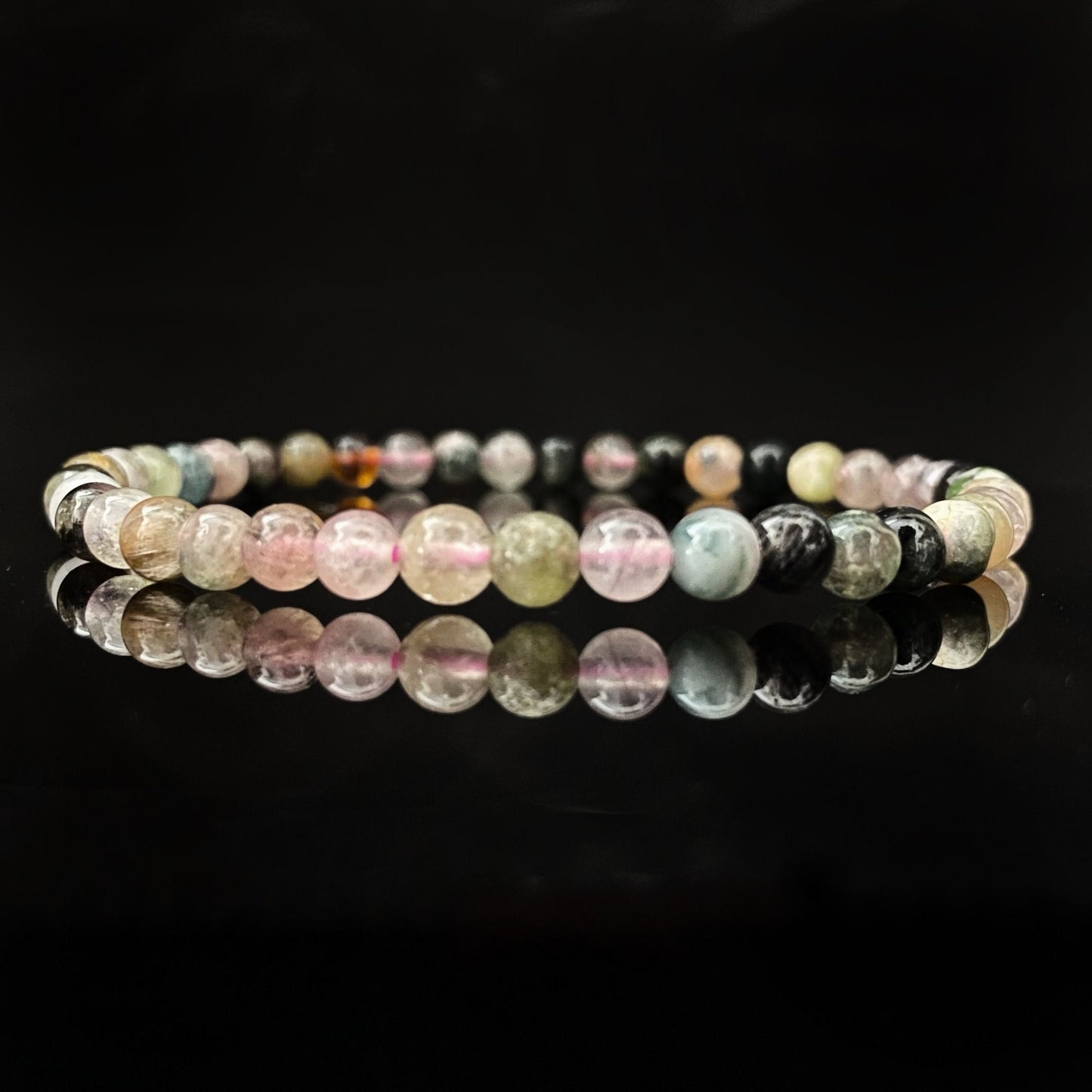 A delicate beaded 4mm Rainbow Tourmaline Bracelet with spherical beads in various shades of pastel colors, including pink, green, and blue, lies on a reflective black surface. The reflection of the bracelet is clearly visible, creating a symmetrical effect and enhancing its chakra balancing properties.