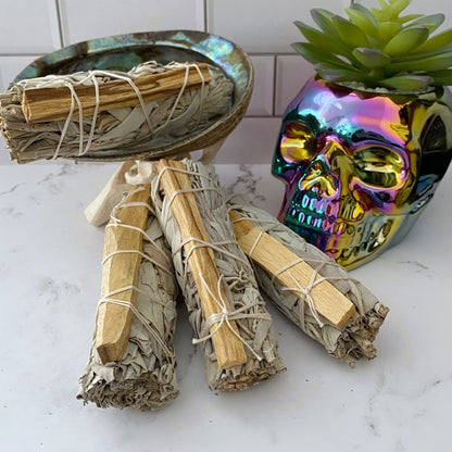 A bundle of Organic White Sage & Palo Santo Smudge Sticks is neatly stacked in front of a shiny, iridescent skull-shaped planter holding a small succulent. The background features a white tiled wall, creating a contrast with the natural and reflective elements in the foreground.