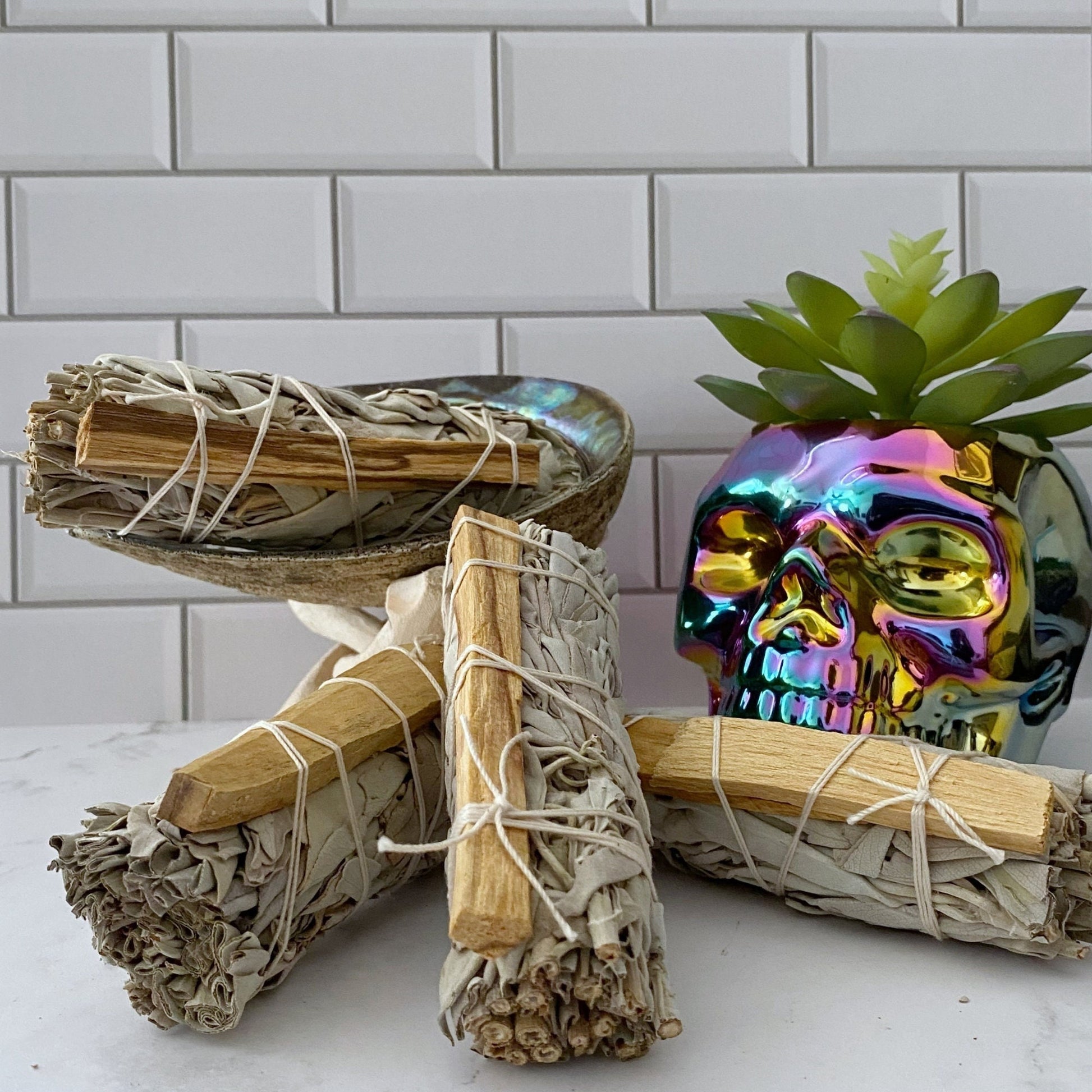 A bundle of Organic White Sage & Palo Santo Smudge Sticks is neatly stacked in front of a shiny, iridescent skull-shaped planter holding a small succulent. The background features a white tiled wall, creating a contrast with the natural and reflective elements in the foreground.