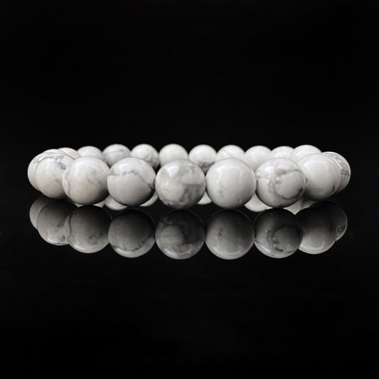 A stretchable 8mm Howlite Beaded Bracelet made of smooth, round white stones with light gray marbling, arranged in a circular pattern against a black background. The bracelet and its reflections create a symmetrical, elegant appearance that embodies the properties of an emotional healing stone.