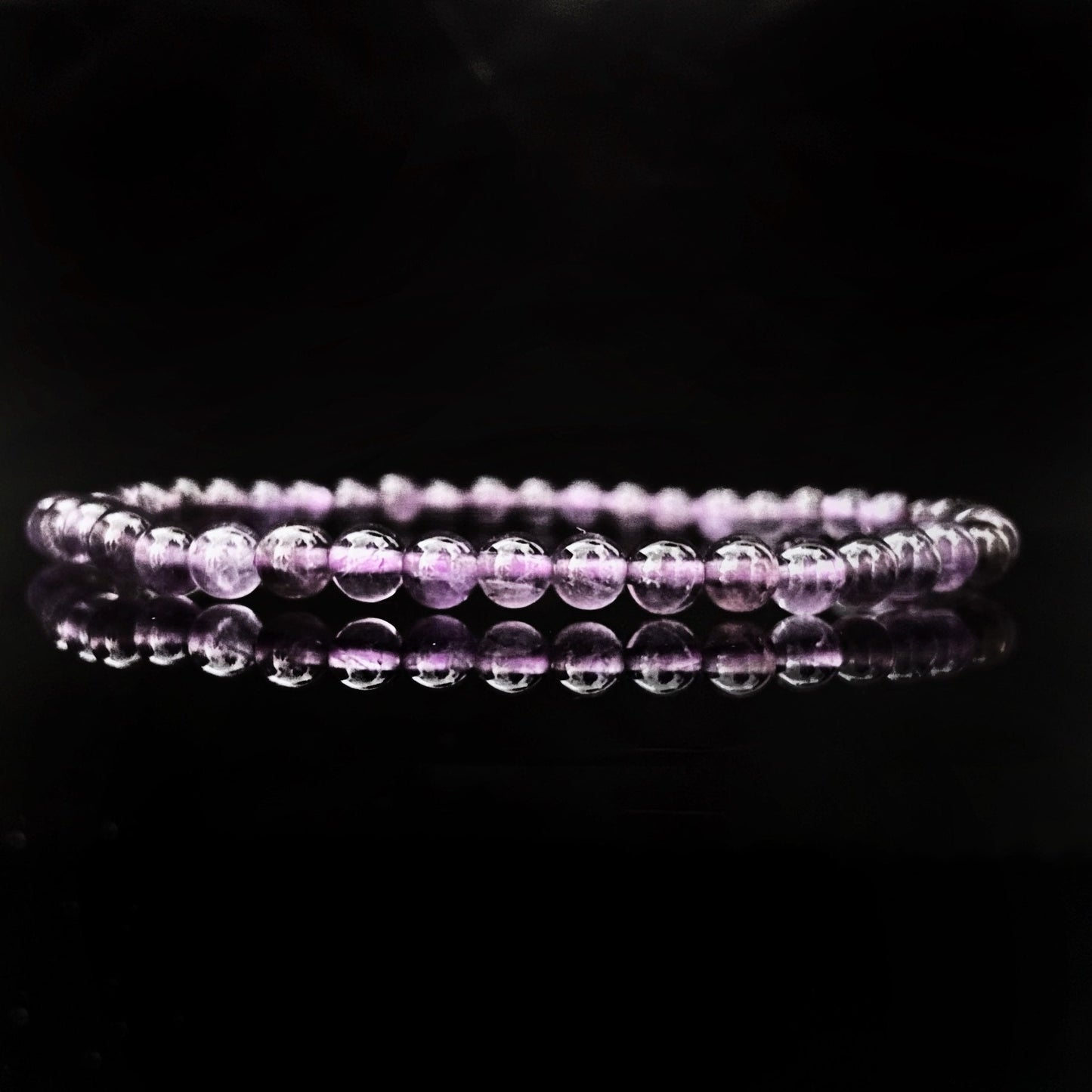 A close-up of a 4mm Amethyst Beaded Bracelet consisting of small, round, translucent purple beads, arranged in a circular pattern and placed against a dark background. The reflection of the bracelet is visible on the glossy surface below it.