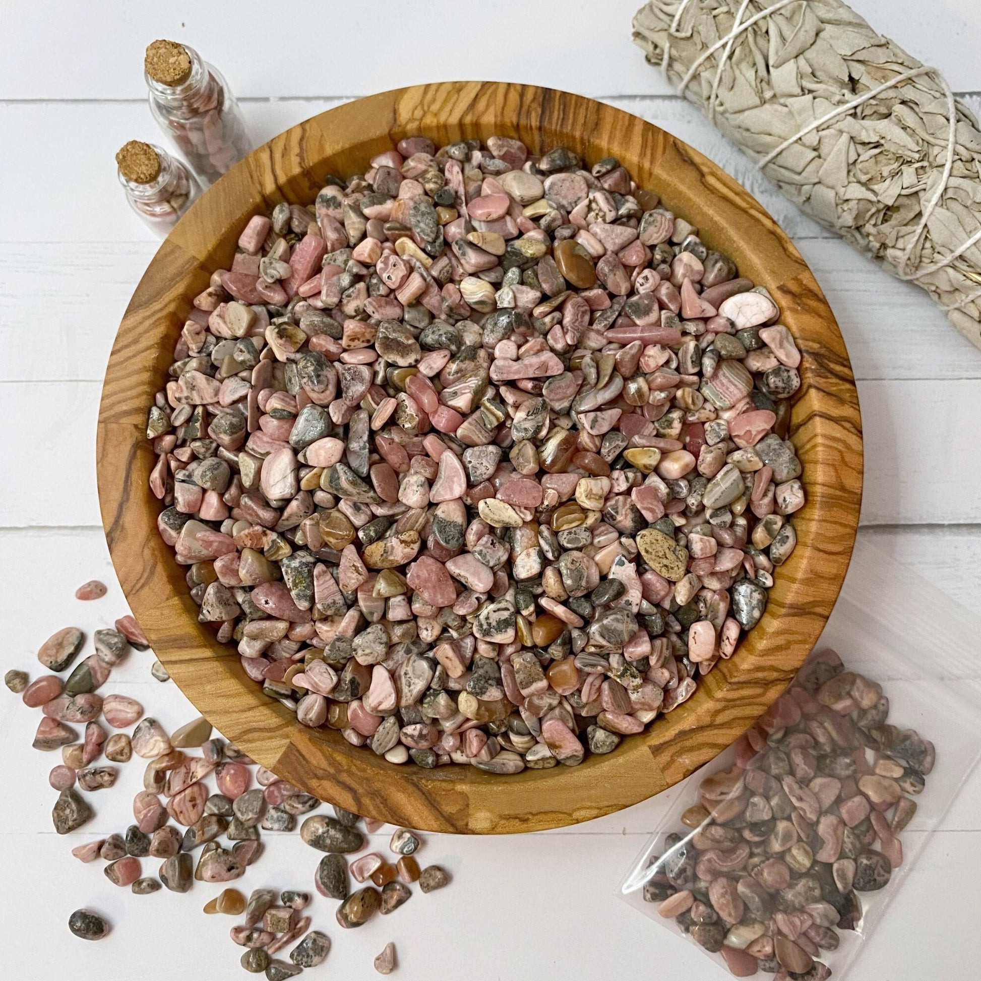 A wooden bowl filled with small polished pink and gray stones, including Rhodochrosite Crystal Chips, sits atop a white wooden surface. Nearby, there is a bundle of sage, a ziplock bag with more stones, scattered stones, and two small glass jars containing stones and crystals—perfect for crafting supplies or metaphysical uses.