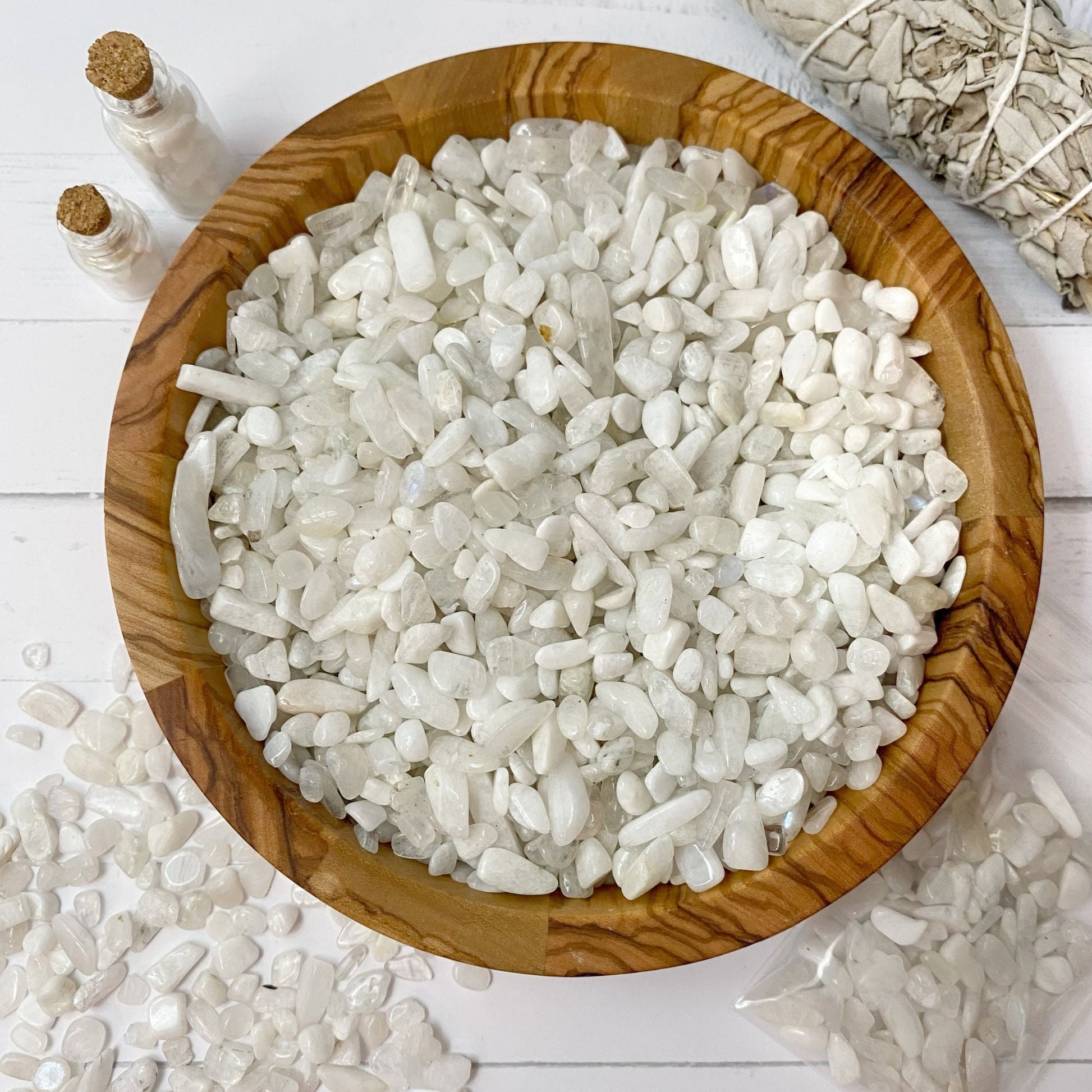 A wooden bowl filled with polished white stones sits on a white surface. Two small vials with cork stoppers and some scattered Rainbow Moonstone Crystal Chips are on the left, while a plastic bag with more stones is partially visible in the bottom right corner. A bundle of sage for metaphysical uses is at the top right.