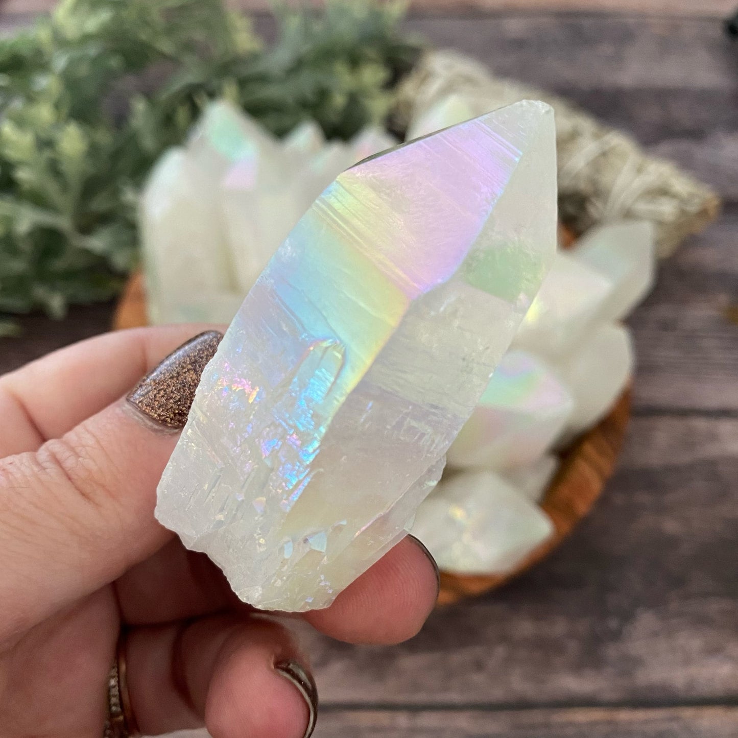 A hand with glittery nail polish holds a large, iridescent 1-4" Raw Angel Aura Quartz Point with a translucent appearance, reflecting rainbow hues. In the background, more similar crystals are displayed in a wooden bowl on a wooden surface, with green foliage partially visible.