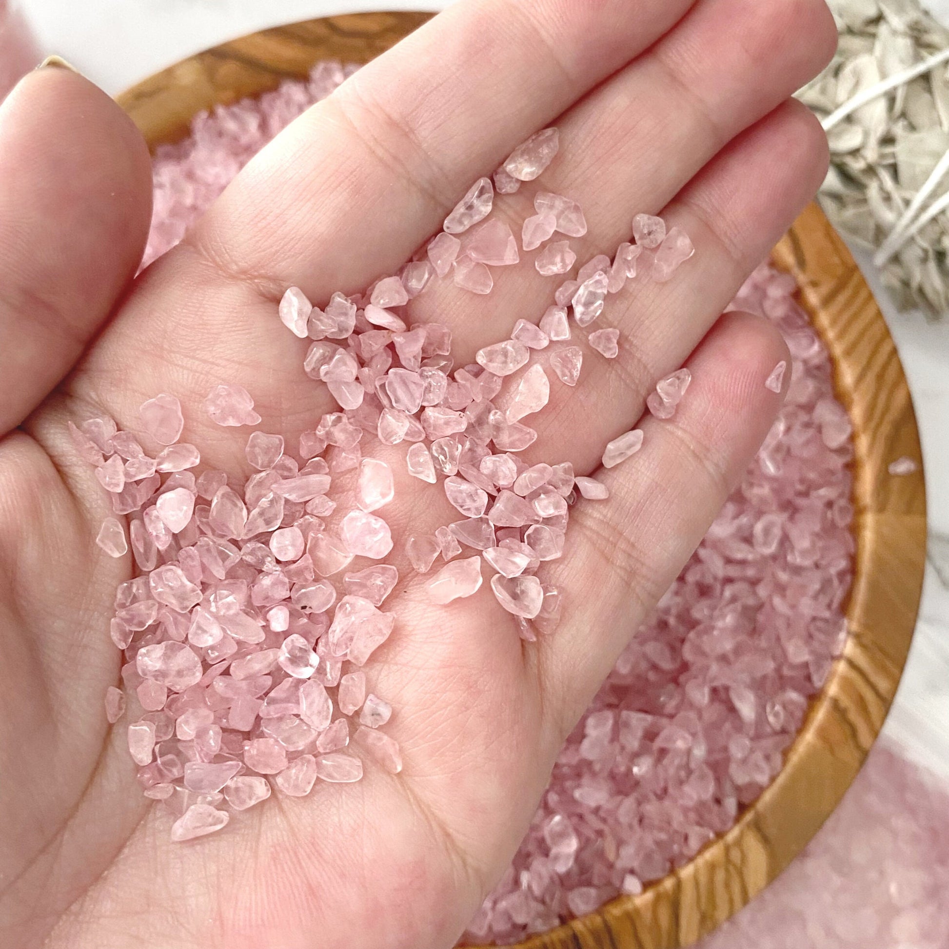 A wooden bowl filled with Rose Quartz Crystal Chips sits on a marble surface. Nearby, two small glass vials with pink salt and a plastic bag containing more crystals accompany a bundle of dried sage wrapped with string. Some pink salt is scattered, mingling gracefully with Rose Quartz Crystal Chips on the surface.