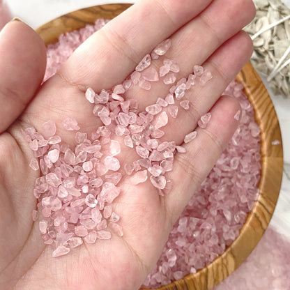 A wooden bowl filled with Rose Quartz Crystal Chips sits on a marble surface. Nearby, two small glass vials with pink salt and a plastic bag containing more crystals accompany a bundle of dried sage wrapped with string. Some pink salt is scattered, mingling gracefully with Rose Quartz Crystal Chips on the surface.