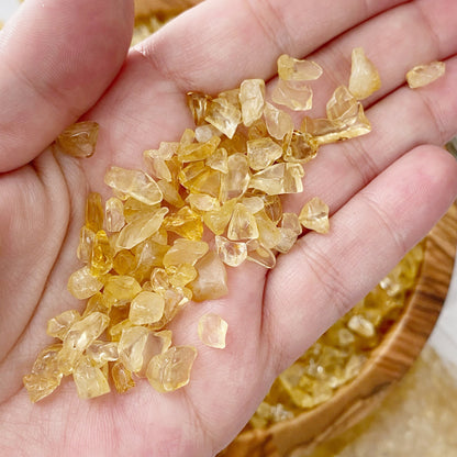 A wooden bowl filled with small, yellow Citrine Crystal Chips sits on a white surface. Surrounding the bowl are a small bundle of sage, two tiny glass bottles filled with similar crystals, and a small plastic bag containing additional stones.