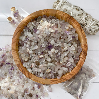 A wooden bowl filled with small, polished mixed crystals, including Super Seven Crystal Chips, is placed on a white surface. Nearby, some tumbled and polished stones are scattered. There are also two small jars filled with crystals and a sage bundle. A plastic bag with additional crystals is also visible.