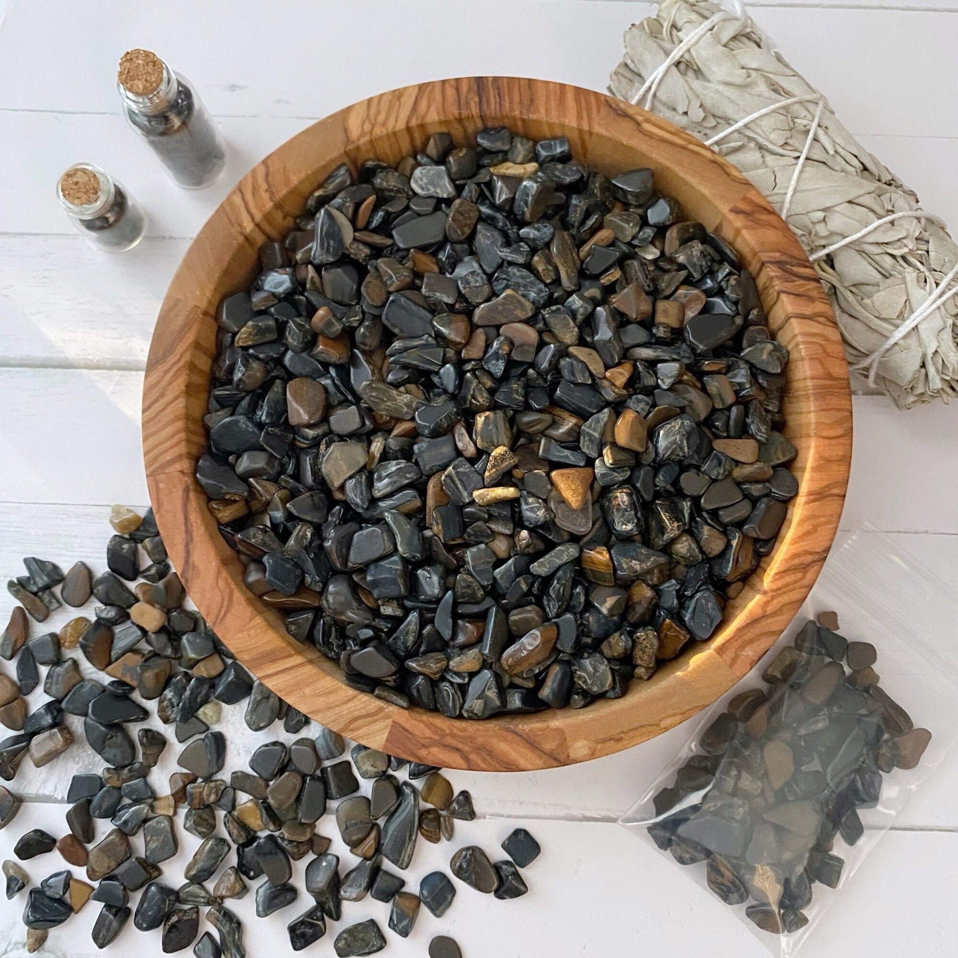 A wooden bowl filled with small, polished black and brown stones, including Blue Tigers Eye Crystal Chips, is placed on a white surface. Some stones are scattered around the bowl. Two small glass bottles with cork stoppers and a bundle of white sage wrapped with string are also visible, hinting at their metaphysical uses.