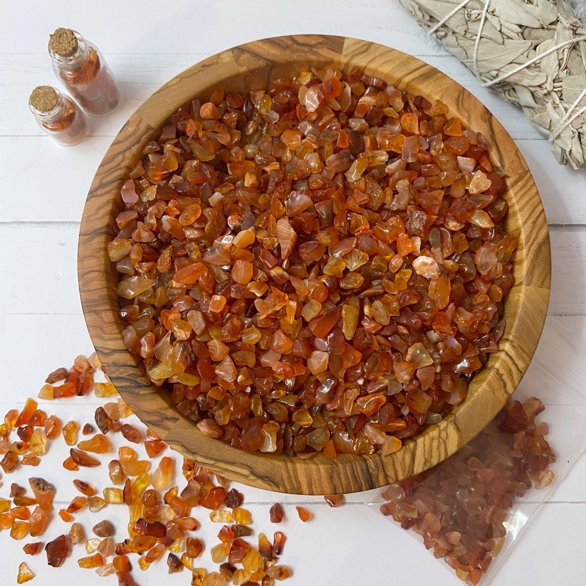 A wooden bowl filled with small, polished Carnelian Crystal Chips in shades of orange and red is centered on a white surface. A smudge bundle, two small vials of stones, and a scatter of additional Carnelian Crystal Chips surround the bowl, perfect for creative and mystical endeavors.