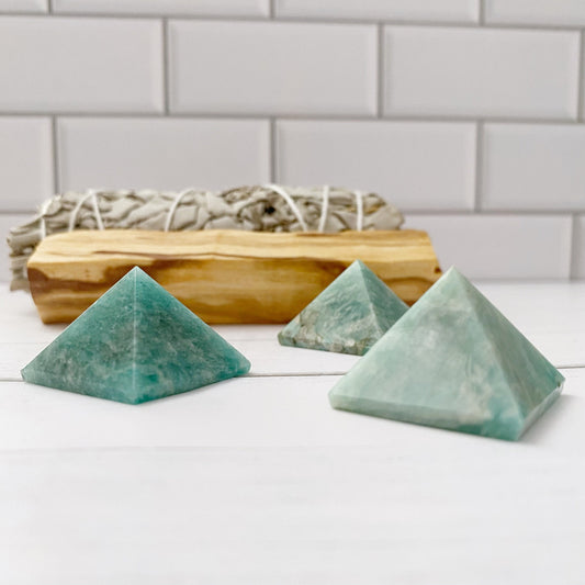 A photo features three 1" Mini Amazonite Pyramids on a white surface, known for stress relief and combating electromagnetic pollution. Behind the pyramids, there is a bundle of dry, white sage sticks. The background consists of white tiled walls, adding a clean and minimalist look to the setup.