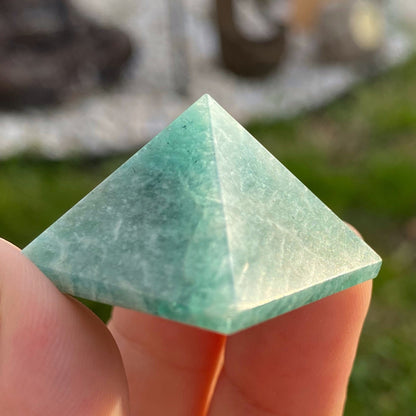 A photo features three 1" Mini Amazonite Pyramids on a white surface, known for stress relief and combating electromagnetic pollution. Behind the pyramids, there is a bundle of dry, white sage sticks. The background consists of white tiled walls, adding a clean and minimalist look to the setup.