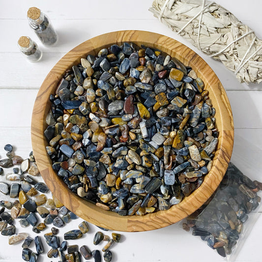 A wooden bowl filled with polished, multicolored tumbled stones is placed on a white surface. Some Pietersite Crystal Chips and other stones are scattered around the bowl. To the right, there is a bundle of sage, a small clear plastic bag with more Pietersite Crystal Chips, and two small glass jars with cork stoppers for metaphysical crafting.