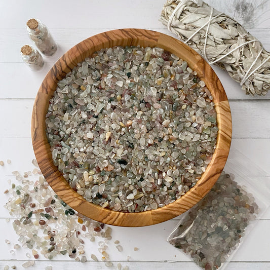 A wooden bowl filled with various small, polished stones sits on a white surface. To the left, some stones are scattered. Two small glass spell jars filled with stones and a bundle of tied sage are positioned nearby. A small bag of polished Mixed Agate Crystal Chips is also visible.