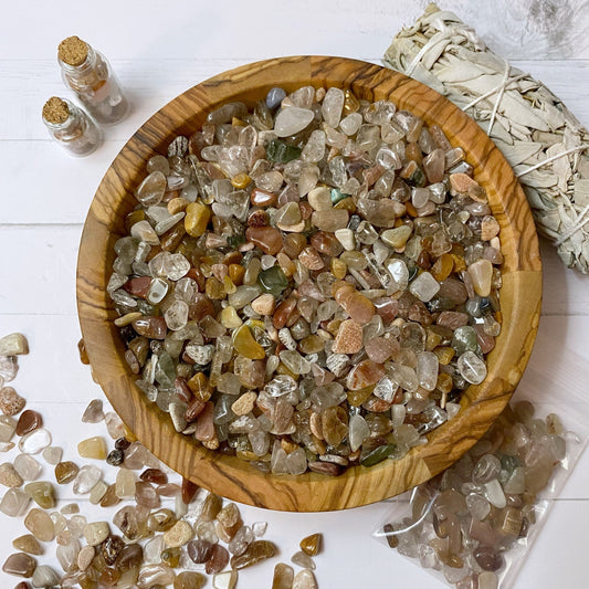 A wooden bowl filled with polished gemstones sits on a white surface. Surrounding the bowl are vials of Rutilated Quartz Crystal Chips, scattered gems, a small sage bundle, and a plastic bag filled with Rutilated Quartz. The scene has a serene and earthy vibe, hinting at its metaphysical uses.