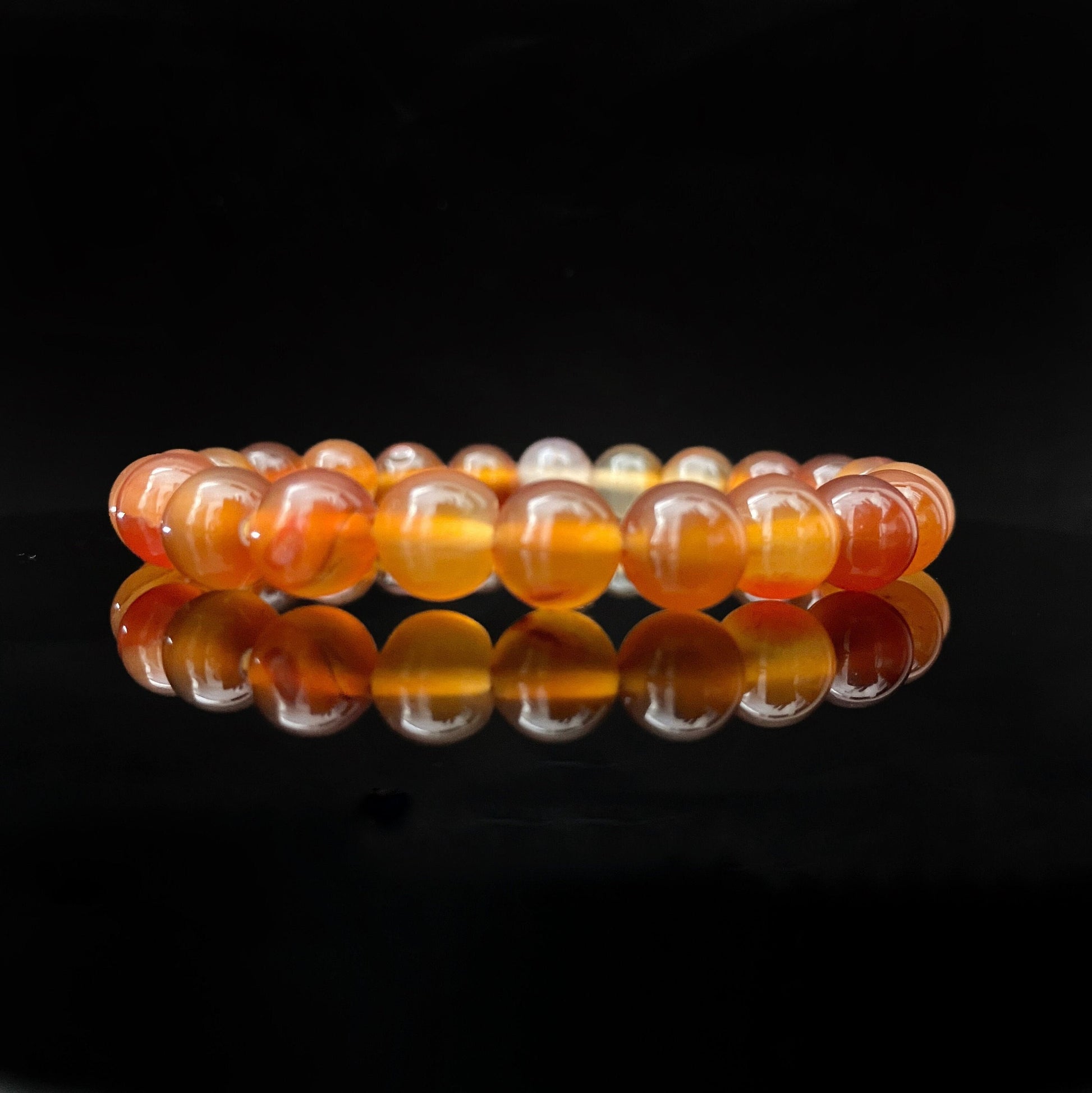 A round 8mm Carnelian Agate Beaded Bracelet made from translucent orange stones is placed on a reflective black surface, creating a mirrored effect. This fertility crystal is composed of smooth, polished beads, varying in shades of orange and yellow.