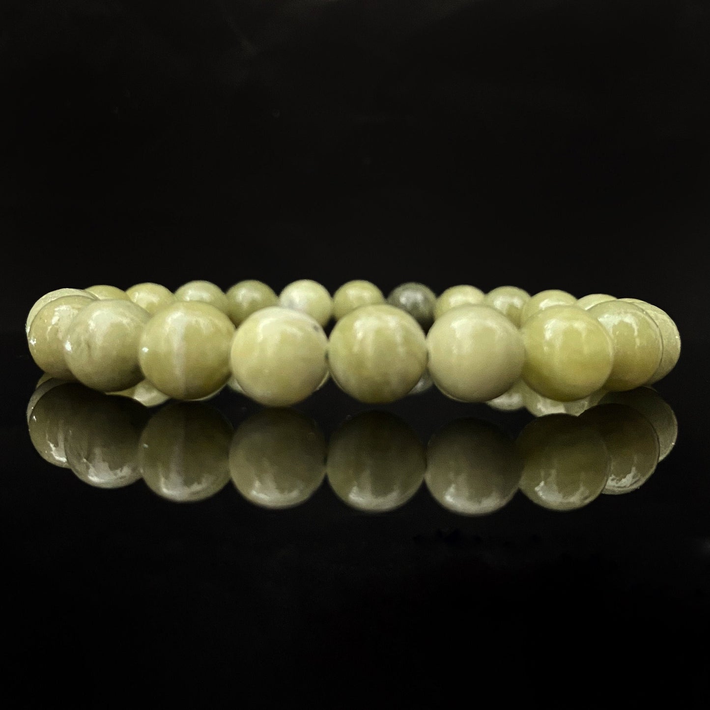 A polished 8mm Chinese Jade Beaded Bracelet forms a closed loop against a solid black background. The smooth, round jade polished beads are a pale green color, and the bracelet's reflection is clearly visible on the shiny surface below.