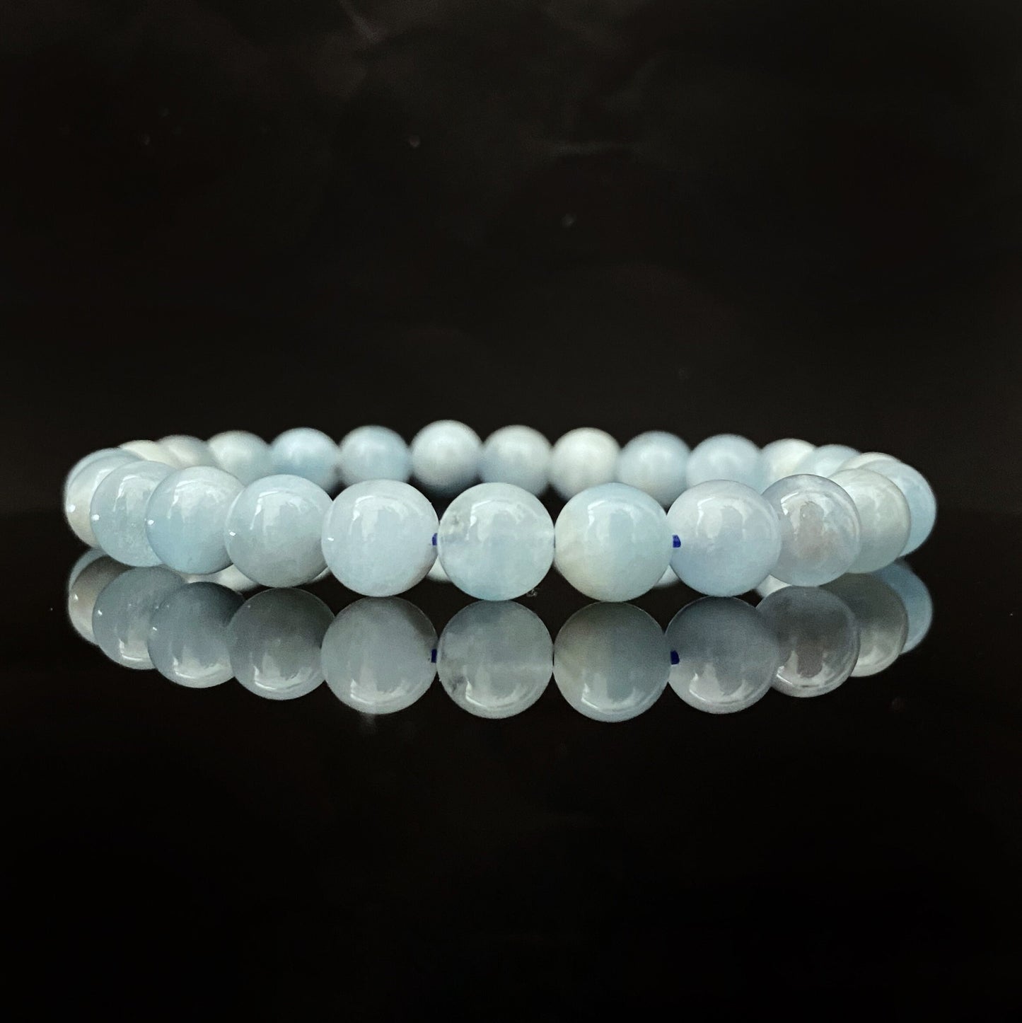 A light blue 8mm Aquamarine Beaded Bracelet is neatly arranged in a circle on a reflective black surface, showcasing its simple, elegant design. Each bead is evenly sized, adding to the harmonious appearance of the accessory, perfect for enhancing your meditation practice.