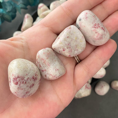 A black bowl filled with smooth, white Cinnabar Tumbled Stones that have pink speckles—reminiscent of Cinnabar crystals—sits on a gray surface. To the left of the bowl is a sprig of red berries with green leaves.