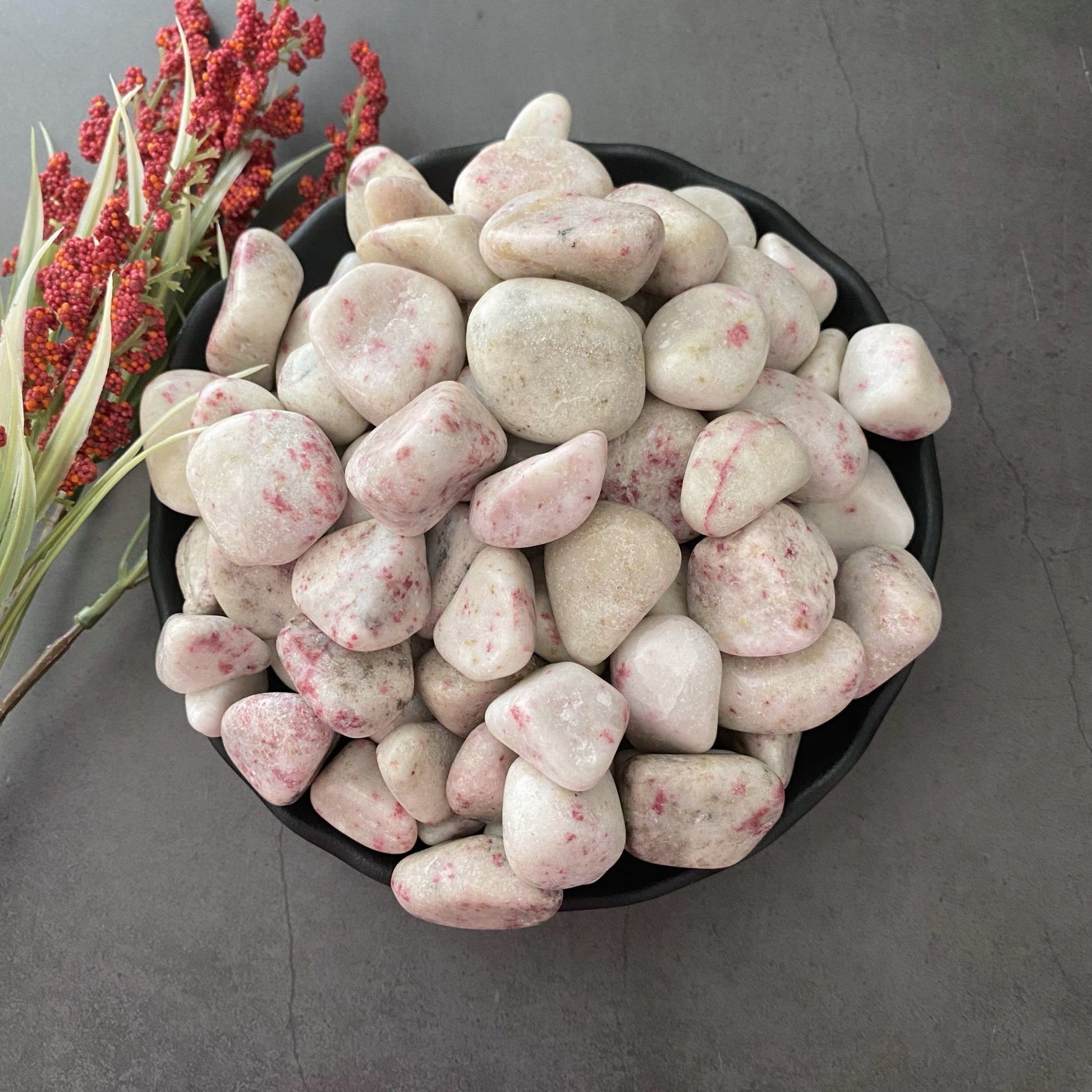 A black bowl filled with smooth, white Cinnabar Tumbled Stones that have pink speckles—reminiscent of Cinnabar crystals—sits on a gray surface. To the left of the bowl is a sprig of red berries with green leaves.