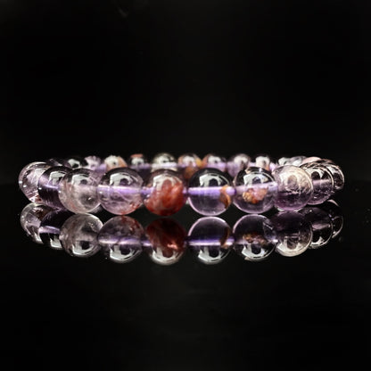 A circular 8mm Super Seven Beaded Bracelet displayed on a glossy black surface, with each bead featuring a mix of translucent purple and reddish hues. The bracelet's reflection is visible below on the polished surface, enhancing its vibrant colors.