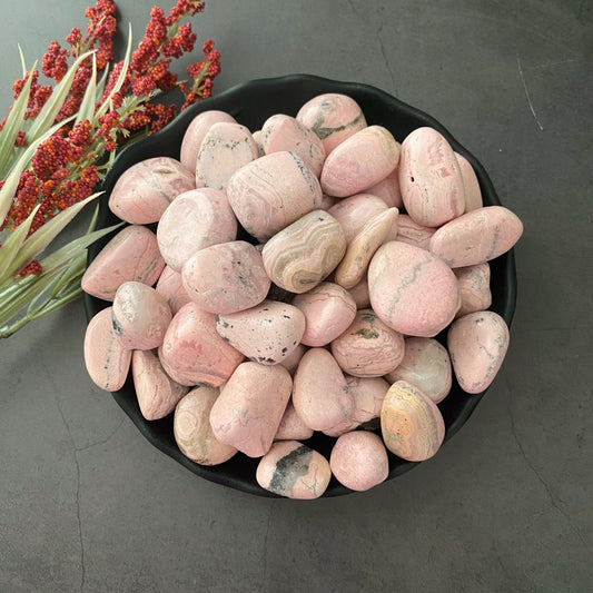 A black bowl filled with smooth, round Rhodochrosite Tumbled Stones is placed on a dark surface. To the left of the bowl, there is a spray of red and green foliage, adding a touch of natural contrast to the scene, ideal for promoting emotional healing.