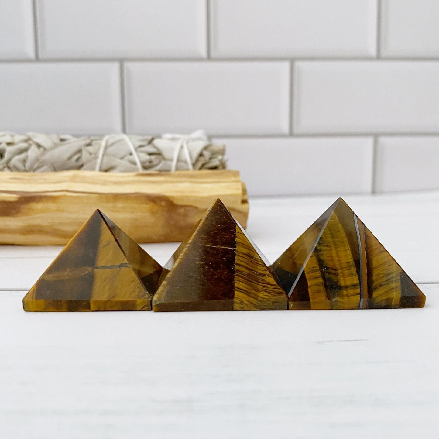 Three small 1" Mini Tigers Eye Pyramids are displayed on a white surface against a white subway tile background. Behind the pyramids, there is a bundle of sage and a piece of Palo Santo wood, enhancing their aura of spiritual awareness.