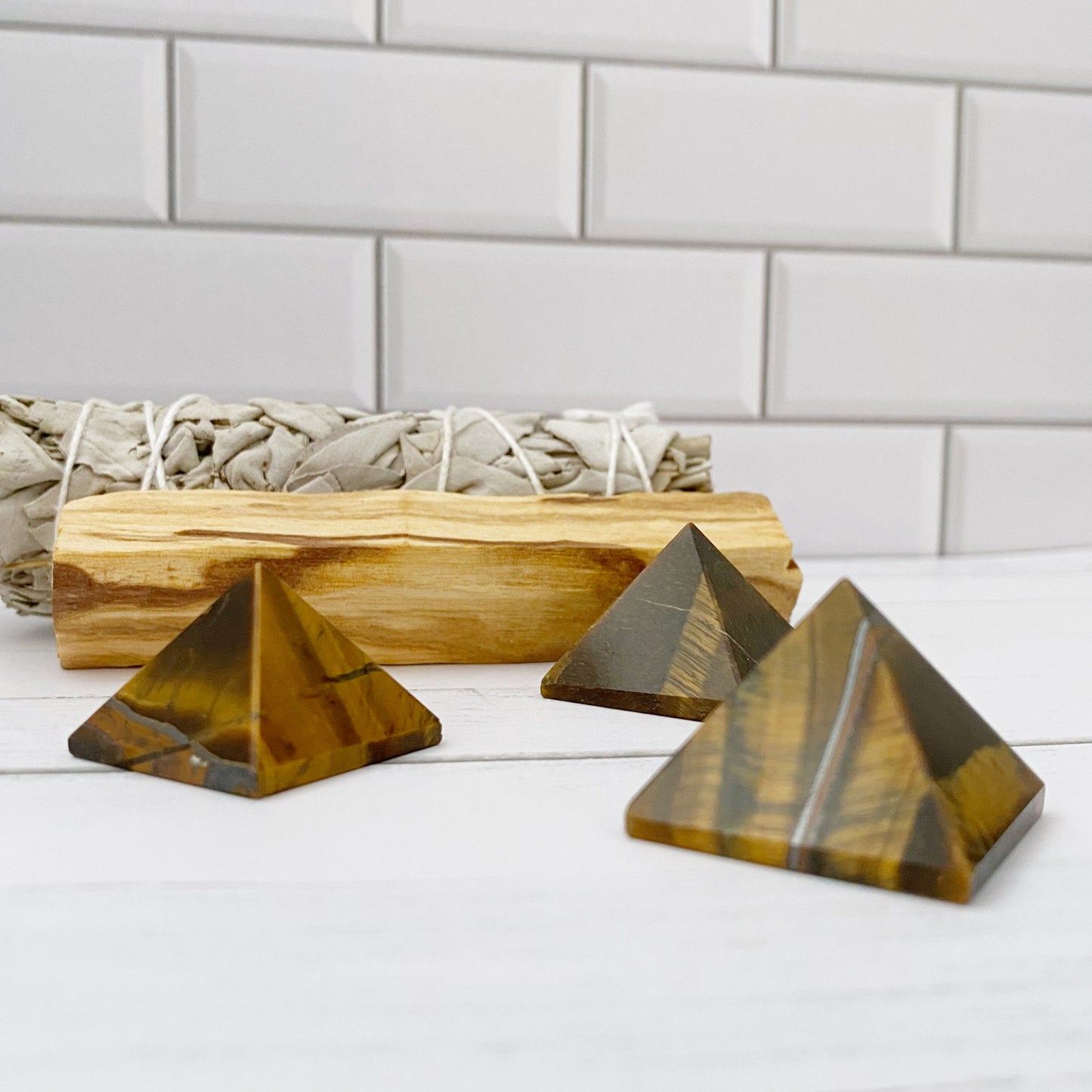 Three small 1" Mini Tigers Eye Pyramids are displayed on a white surface against a white subway tile background. Behind the pyramids, there is a bundle of sage and a piece of Palo Santo wood, enhancing their aura of spiritual awareness.